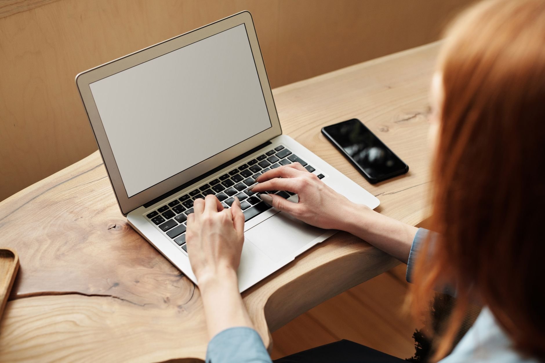 A woman working on a laptop at a wooden desk, indoors with natural light.